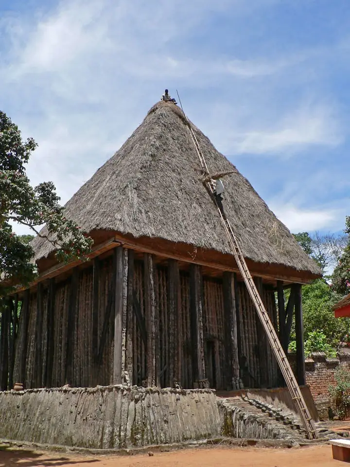 A tall thatched building standing on close-set carved wooden pillars, against an open sky.