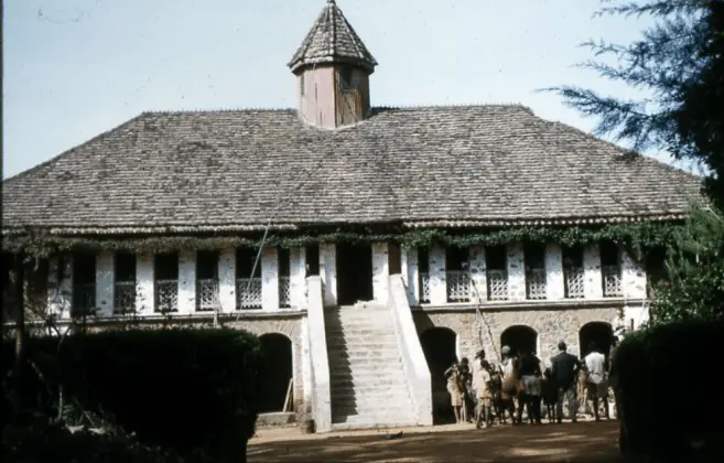 An older photograph of a long shingled roof with a lantern tower, people climbing the front staircase.